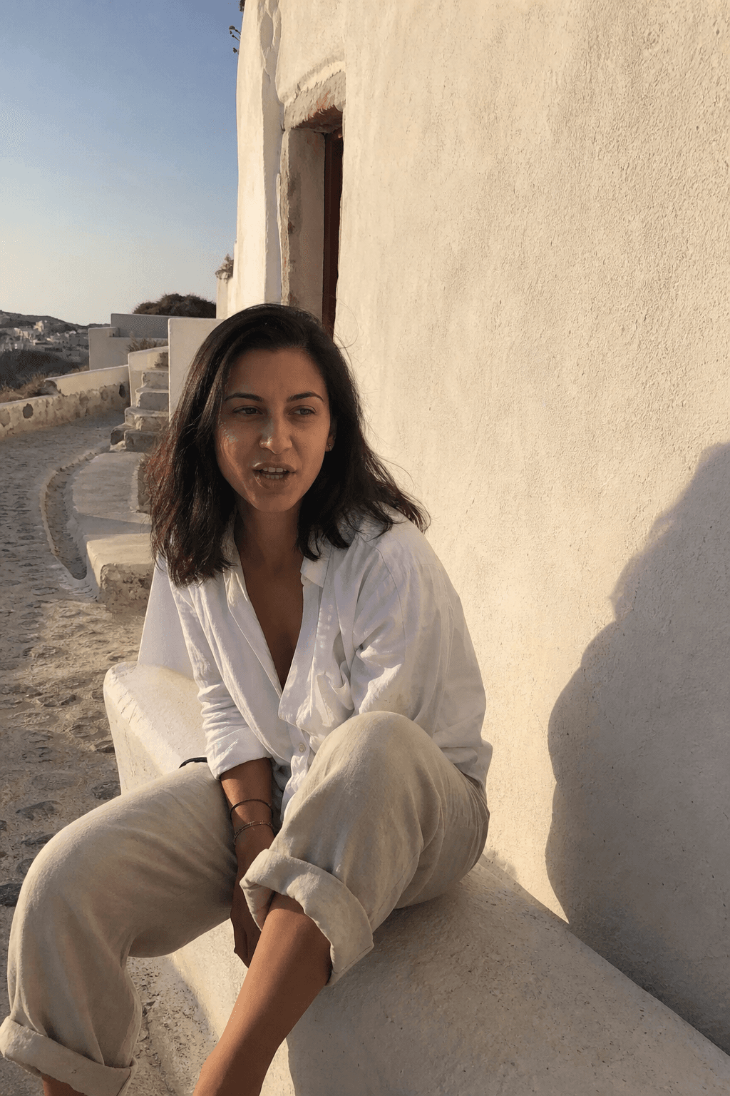 Assise sur un muret blanc à Oia, en chemise de lin, tôt le matin.